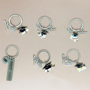Op de afbeelding: Een verzameling sleutelhangers met een afstudeerthema. Elke sleutelhanger heeft een zilveren ring met bedels, waaronder een afstudeerhoed, een diploma en het jaar 2026. Eén sleutelhanger heeft een label met de tekst "I GRADUATED!" en een citaat.
