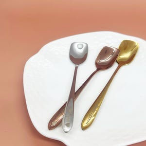 May include: Three metal spoons with square bowls rest on a white, scalloped plate. One spoon is silver, one is rose gold, and one is gold. The rose gold spoon has a heart design, and the gold spoon has the words "Baby Spoon".