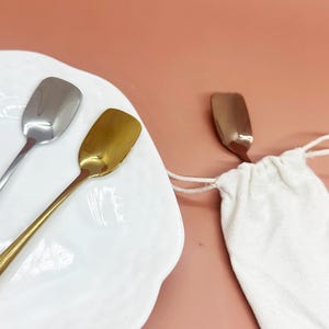 May include: Three small spoons with rectangular bowls. One spoon is silver, one is gold, and the third is copper-colored. The spoons are on a white plate and next to a small white drawstring bag. The background is a solid peach color.