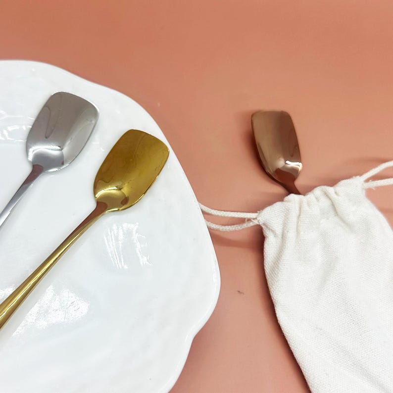 May include: Three small spoons with rectangular bowls. One spoon is silver, one is gold, and the third is copper-colored. The spoons are on a white plate and next to a small white drawstring bag. The background is a solid peach color.
