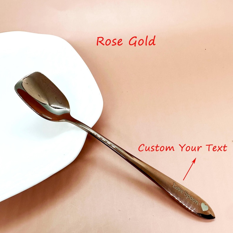 May include: Rose gold spoon with a heart-shaped detail and the text "Love Spoon". The spoon is resting on a white plate, with the words "Custom Your Text" and "Rose Gold" in the background.