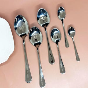 May include: A selection of stainless steel spoons, each engraved with a different cartoon face and text. The spoons are of varying sizes. Text includes "Grandpa fun spoon", "The humorous spoon", and "King smiling spoon".