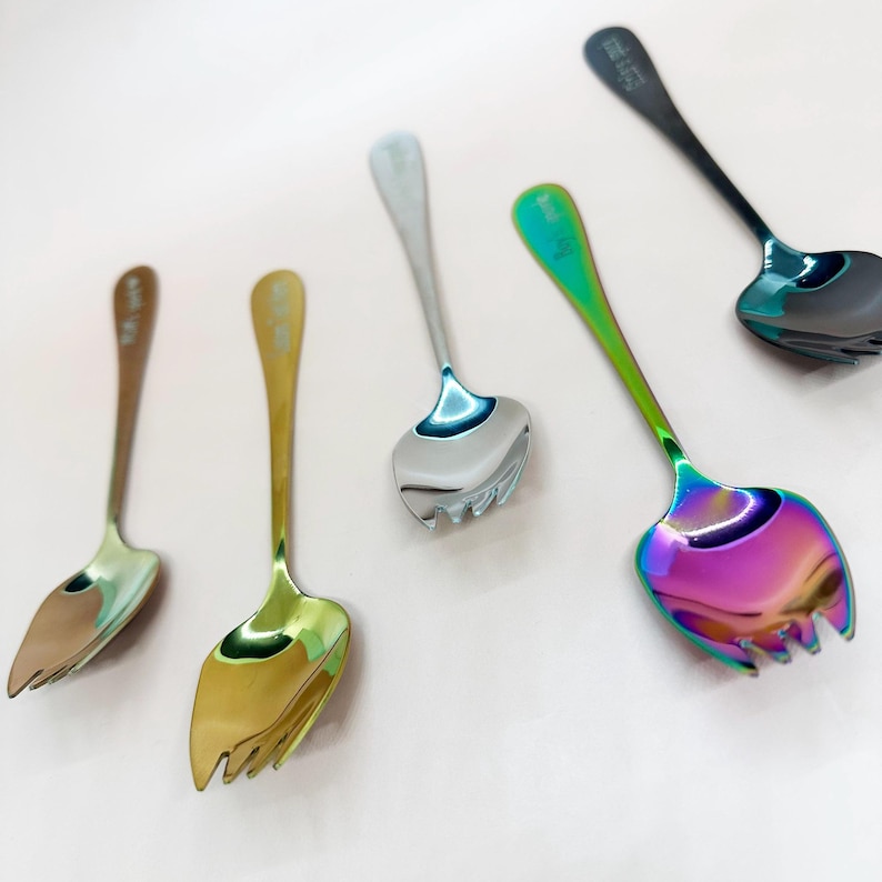 May include: A collection of serving utensils with a distinctive design. The utensils have a fork-like end and a spoon-like bowl. The colours include gold, silver, bronze, and a rainbow iridescent finish. The utensils are arranged on a light pink surface.