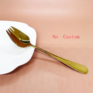 May include: A gold-coloured serving fork with a three-pronged head and a long handle. The fork is on a white, irregularly shaped plate. The words "No Custom" are in red.