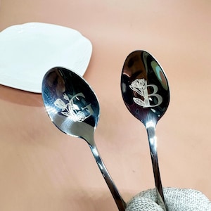 May include: Two stainless steel spoons with engraved floral designs and initials. One spoon features the letter "C" and the other the letter "B". A white, scalloped plate is in the background.