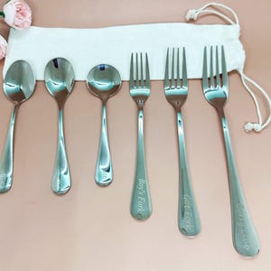 May include: A set of six stainless steel spoons and forks, including three spoons and three forks, with engraved text on the handles. The set is displayed on a pink surface with a white drawstring bag in the background.