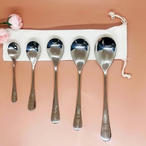 May include: Five stainless steel spoons of varying sizes are displayed against a white cloth, with pink flowers in the upper left corner. Each spoon has an engraved inscription: "child spoon", "Boy's Spoon", "Party Gift", "Tom's Spoon", and "Spoon for Jack".