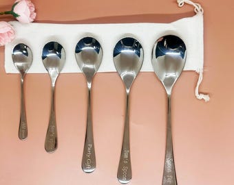 May include: Five stainless steel spoons of different sizes are arranged against a white cloth, with pink flowers in the upper left. Each spoon is engraved with a name or phrase: "child spoon", "Boy's Spoon", "Party Gift", "Tom's Spoon", and "Spoon for Jack".