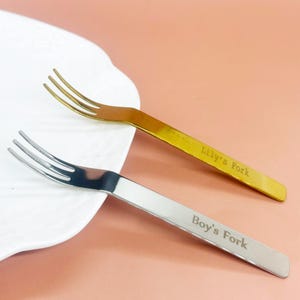 May include: Two small forks, one gold and one silver, are displayed on a white plate and a pink surface. The gold fork is engraved with "Lily's Fork," and the silver fork reads "Boy's Fork." Each fork has three tines.
