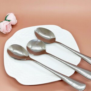 May include: Three shiny, stainless steel soup spoons arranged on a white, decorative plate. The spoons have long, slender handles and rounded bowls. The handles are engraved with the words "Early Eye". Pink roses are in the background.