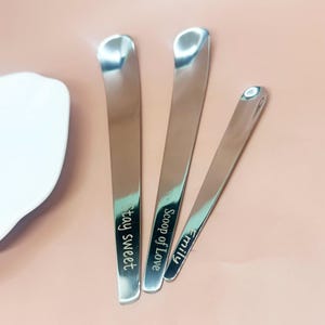 May include: Three stainless steel serving spoons with curved handles. One spoon reads "stay sweet", another reads "Scoop of Love", and the third reads "Emily". The spoons are arranged on a pink surface.
