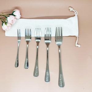 以下が含まれることがあります： さまざまなサイズのステンレス鋼のフォーク6本セット。それぞれに異なる名前が刻印されています。「Small Fork」、「Girl's Fork」、「Boy's Fork」、「Gift Fork」、および「Curry Club」。フォークは、白い布のバッグとピンクの花が添えられた、明るいピンク色の表面に表示されています。