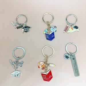 May include: A selection of graduation keyrings. Each keyring has a silver ring with charms such as graduation caps, books, pencils, and the text "2026 Complete Grad" and "Graduation is not the end, it's the beginning."