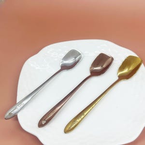 Puede incluir: Tres cucharas con cuencos cuadrados y mangos largos descansan sobre un plato blanco. Las cucharas son plateadas, bronce y doradas. Cada cuchara tiene texto y un detalle en forma de corazón en el mango.
