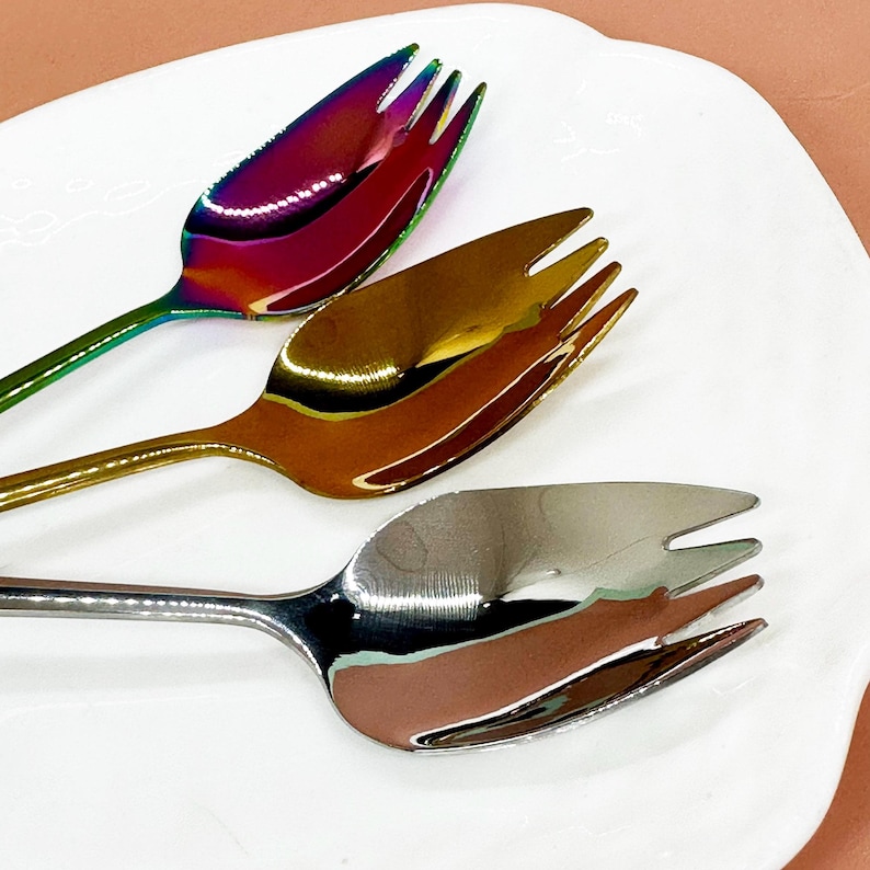 May include: Three decorative forks with a unique design. The forks have a metallic finish in iridescent, gold, and silver colours. The forks are placed on a white, oval-shaped plate. The forks are designed for serving appetizers or desserts.