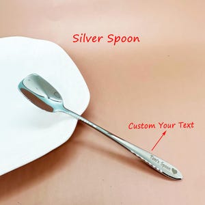May include: A shiny silver spoon with a heart symbol and the text "Tom's Spoon" engraved on the handle. The spoon is next to a white plate. The text "Silver Spoon" and "Custom Your Text" are also visible.