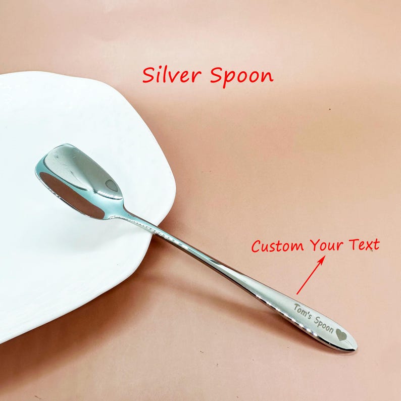 May include: A shiny silver spoon with a heart symbol and the text "Tom's Spoon" engraved on the handle. The spoon is next to a white plate. The text "Silver Spoon" and "Custom Your Text" are also visible.