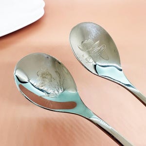 Puede incluir: Dos cucharas de acero inoxidable con diseños florales grabados. Las cucharas tienen un acabado pulido y una forma de cuenco curvada. El diseño floral parece ser una flor de loto. Las cucharas están sobre una superficie rosa.