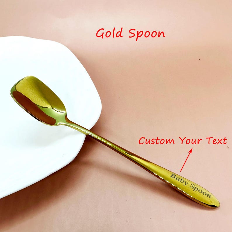 May include: A gold-colored spoon with a square bowl and a long handle. The text "Baby Spoon" is engraved on the handle. The spoon is next to a white plate on a pink surface. The text "Gold Spoon" and "Custom Your Text" are also visible.