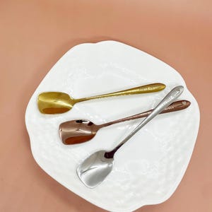 May include: Three small spoons in gold, copper, and silver rest on a white, irregularly shaped plate. The spoons have rectangular bowls and long, slender handles. The gold spoon has the text "Fairy Spoon" on the handle.