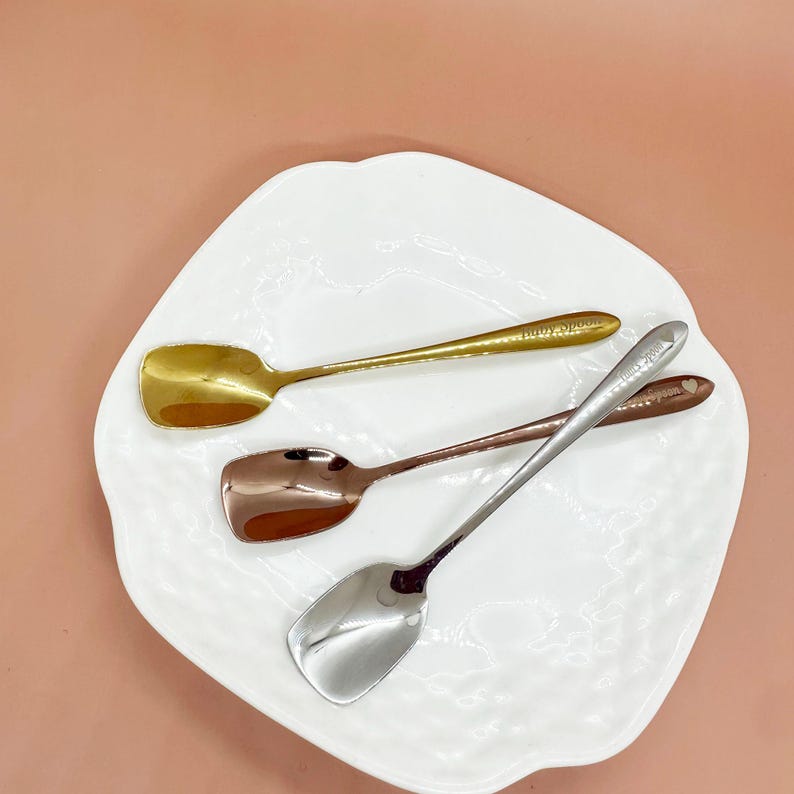 May include: Three small spoons in gold, copper, and silver rest on a white, irregularly shaped plate. The spoons have rectangular bowls and long, slender handles. The gold spoon has the text "Fairy Spoon" on the handle.