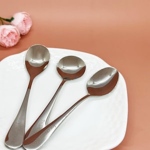 Op de afbeelding: Drie glanzende roestvrijstalen lepels op een wit bord. De lepels hebben lange, slanke stelen en afgeronde kommen. Eén lepel is gemarkeerd met "Boy Spoon", een andere met "Tea" en de derde met "Ice cream Spoon".