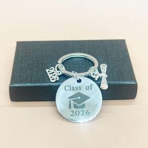 May include: Silver graduation keychain with a round pendant engraved with a graduation cap and the text "Class of 2026". The keychain also features charms with the numbers "2026" and a graduation scroll.