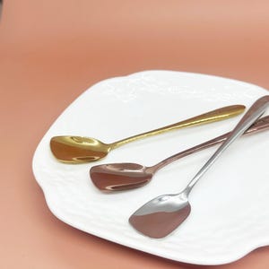 May include: Three spoons with square-shaped bowls rest on a white plate. The spoons are gold, copper, and silver. The spoons are arranged diagonally across the plate, with the gold spoon at the top.
