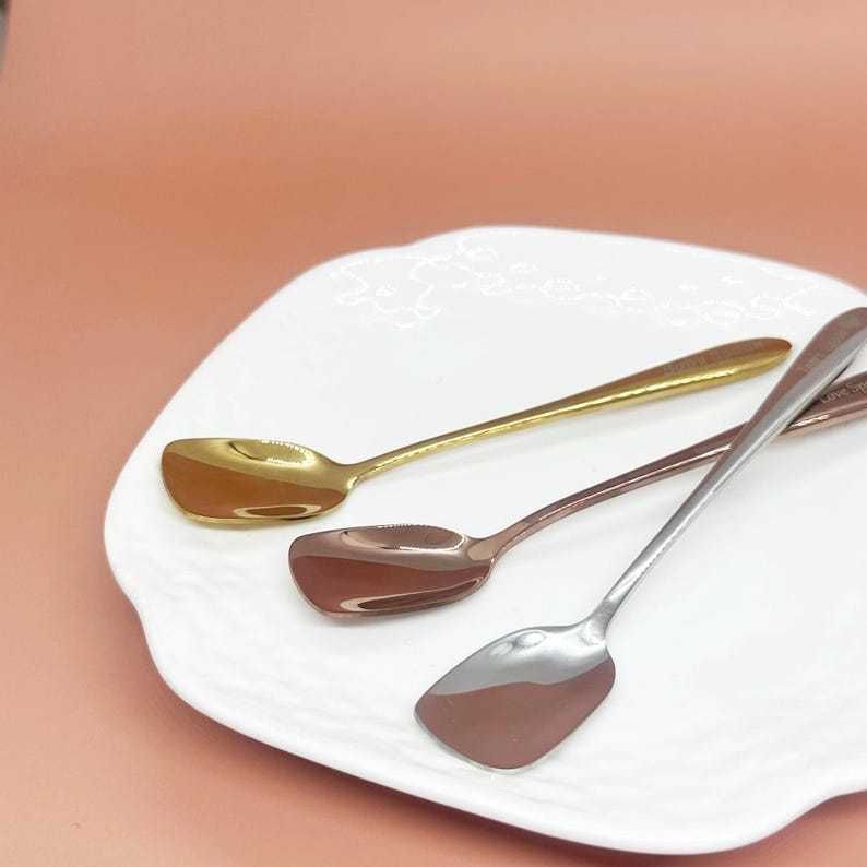 May include: Three spoons with square-shaped bowls rest on a white plate. The spoons are gold, copper, and silver. The spoons are arranged diagonally across the plate, with the gold spoon at the top.