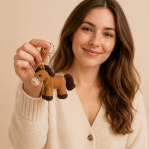 May include: A small, hand-knitted horse keyring featuring a tan body, dark brown mane, tail, and hooves, and a cream-coloured muzzle. The plush horse is attached to a silver-toned metal split ring and chain, designed for use as a decorative accessory.