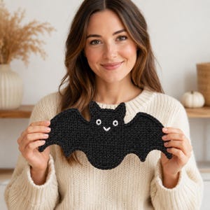May include: A black knitted bat with white embroidered eyes and a smiling mouth. The bat has outstretched wings and pointed ears. The bat is held up in front of a person wearing a cream-colored sweater. The background includes a vase with dried plants and a small white pumpkin.