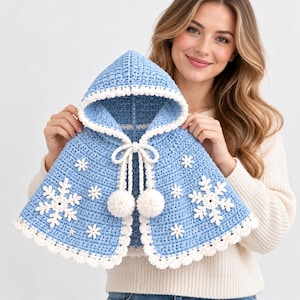 May include: A light blue hooded crochet cape featuring white snowflake appliqués, a white scalloped trim along the edges, and a white drawstring closure finished with two large white pom-poms at the ends of the cords.