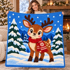 May include: A square chunky knit blanket featuring a central brown deer with large antlers and a red scarf with a white snowflake pattern. The background depicts a snowy winter landscape with dark green pine trees and a bright blue sky.