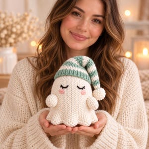 May include: A small, cream-coloured knitted ghost figurine featuring a soft texture, closed embroidered eyes, pink stitched cheeks, and a green and white striped stocking cap topped with a small white pom-pom, held in two hands.