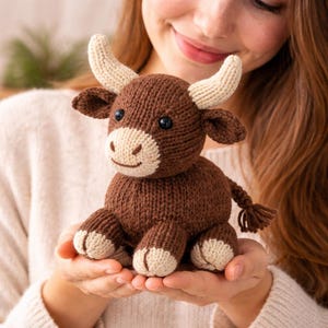 Könnte beinhalten: Handgestricktes Stier-Spielzeug in Braun- und Cremetönen mit schwarzen Augen und cremefarbenen Hörnern. Der Stier sitzt aufrecht, mit einem kleinen Schwanz. Das Spielzeug wird in den Händen gehalten.