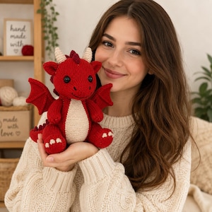 Könnte beinhalten: Ein kleines, handgefertigtes rotes Drachen-Stofftier mit cremefarbenem Bauch, zwei kleinen Hörnern und Flügeln. Der Drache hat schwarze Knopfaugen, ein aufgesticktes Lächeln und kleine cremefarbene Krallen an den Füßen, alles aus Strickgarn.