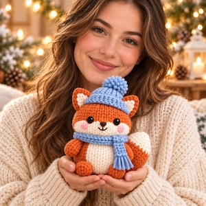 May include: A small, handcrafted crochet fox plush toy featuring orange and cream yarn, wearing a light blue knitted hat with a pom-pom and a matching light blue scarf. The fox has black safety eyes, a stitched nose, and pink blush accents.