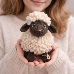 Puede incluir: Un juguete de oveja de ganchillo hecho a mano con un cuerpo esponjoso de color crema, cara, orejas y patas de color marrón oscuro. La oveja tiene ojos grandes y expresivos y una pequeña nariz cosida de color rosa. Una pequeña etiqueta de madera cuelga de su pata.