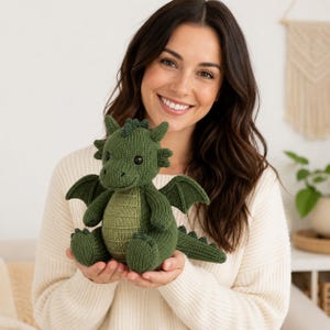 May include: A knitted stuffed dragon toy in a deep forest green color with a lighter sage green belly. The dragon features small wings, a textured spine, and dark round eyes. It is crafted with a visible knit stitch pattern throughout its body.