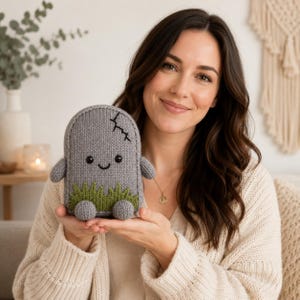 May include: A grey knitted plush toy shaped like a tombstone with a rounded top, featuring a stitched black smile, two small black eyes, a patch of green grass at the base, and a small black stitched crack detail on the upper right side.
