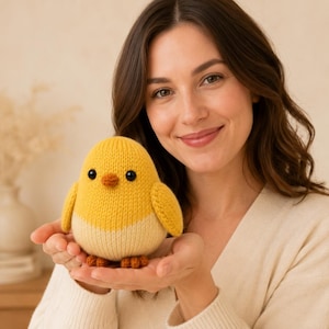 May include: A small, round, knitted chick toy featuring a bright yellow upper body, a cream-coloured lower section, two dark brown button eyes, a small orange beak, and two orange feet. The texture is soft and clearly shows the knitted yarn stitches.