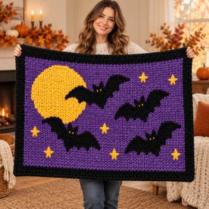 May include: A chunky knit blanket featuring a purple background with a large yellow moon, four black bats, and several small yellow stars. The blanket is framed by a thick black border and displays a textured, hand-knitted appearance.