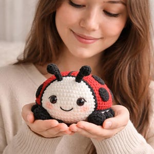 May include: A handmade crochet ladybug plushie with a red and black shell, white head, and black antennae. The plushie has a smiling face and is held in open hands. The ladybug is a cute, soft toy.