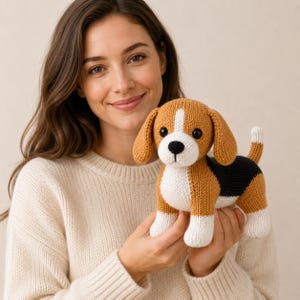 Puede incluir: Un juguete de beagle tejido con un patrón tricolor de hilo marrón, blanco y negro. El perro de peluche tiene orejas caídas, una franja blanca en la cara, una nariz negra y una pequeña cola erguida, todo con una textura de punto visible.