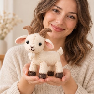 May include: A handcrafted felt goat plush toy featuring a cream-coloured body, dark brown hooves, and curved brown horns. The goat has black bead eyes, a stitched mouth, a small white beard, and pink-lined ears with visible tan blanket stitching.