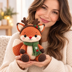 Può includere: Un peluche a forma di volpe lavorato a maglia con filato arancione e crema, che indossa una sciarpa verde con un fiocco di neve bianco e bacche rosse. La volpe ha corna marrone scuro, occhi neri, un naso rosso e zampe marrone scuro.