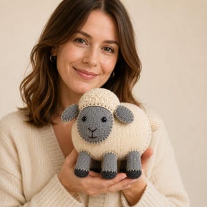 May include: A knitted sheep toy made from fabric, featuring a cream-coloured body and head with a textured top. The face, ears, and legs are dark grey, accented with visible white stitching around the edges. It has two black bead eyes.