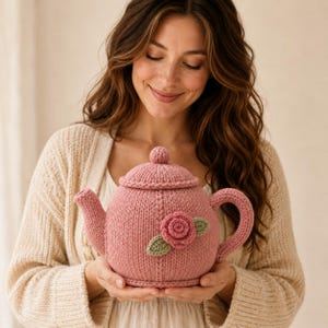 May include: A dusty pink knitted tea cozy featuring a textured surface with a central three-dimensional rose applique in a darker shade of pink, accompanied by two small sage green knitted leaves, a rounded lid, a curved handle, and a spout.