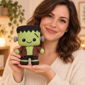 May include: A small, handmade felt plush toy shaped like a green monster with a flat black top, black stitched hair, and two grey bolts on the sides of its head. The figure wears a brown jacket over black trousers and has black eyes and a stitched smile.