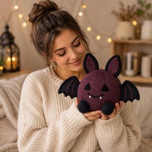 May include: A round, dark purple knitted bat plush toy with black wings, black ears with purple centres, two black triangular eyes, and a small black mouth with two white fangs. The bat has two small black feet at the bottom of its spherical body.
